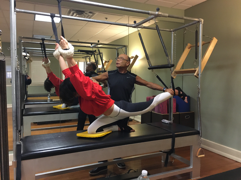 Person exercising with instructor on Pilates reformer machine. Both are in a gym setting.