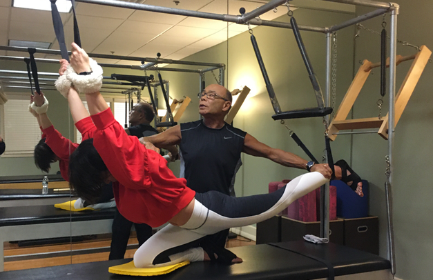 Woman doing Pilates on a specialized apparatus, instructor assisting. Exercise room, white, black, red clothing.