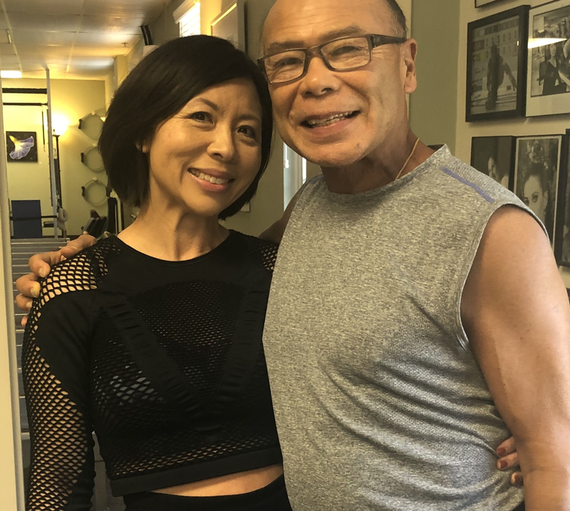 Woman and man smiling, posing indoors. Woman in black top, man in gray tank top, both arm-in-arm.