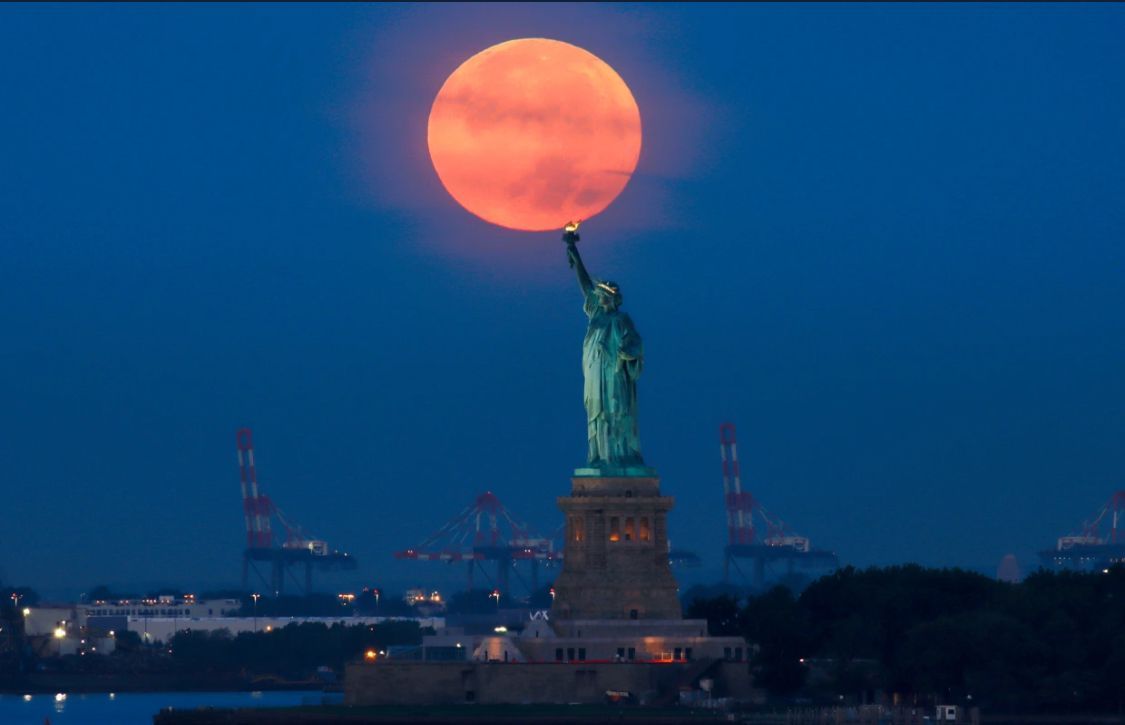 Statue of Liberty holding up a large, orange full moon against a dark blue sky.