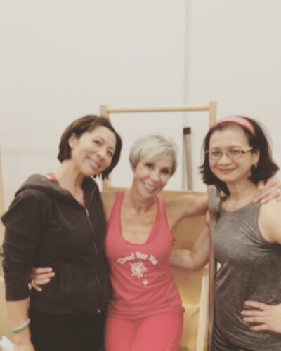 Three women smiling, posing for photo in a studio. The woman in the middle is wearing a pink outfit, arms around the other women.