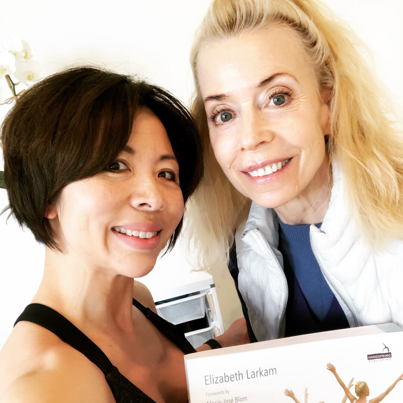 Two women smiling, posing for the camera. One holds a box labeled 