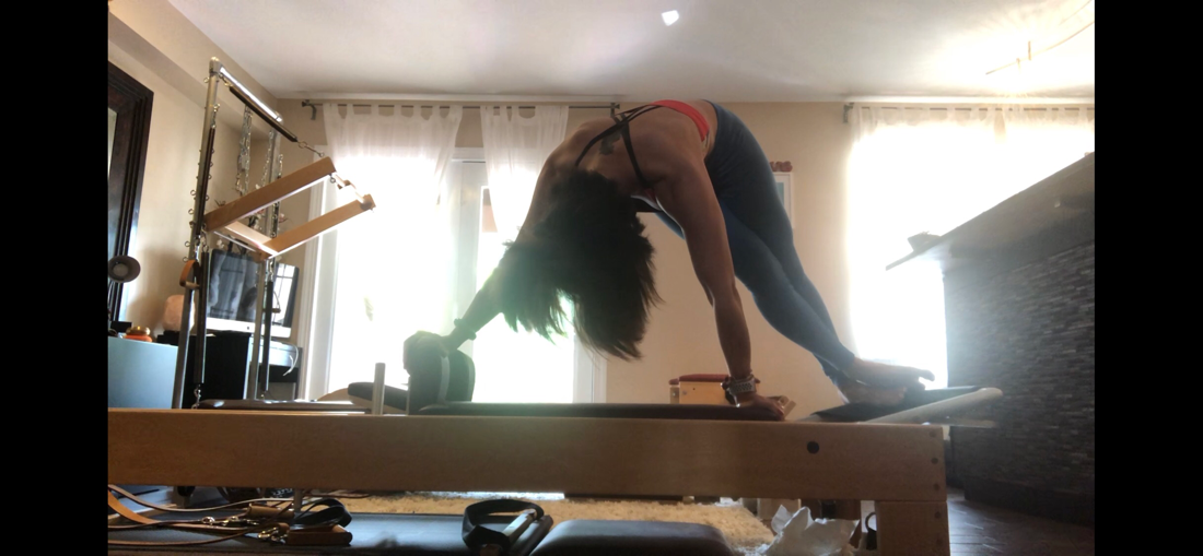 Woman on Pilates reformer, in a back bend. Hands on the frame, feet on foot bar, in a room with blinds.