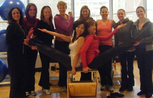 Group of women posing, two sitting on Pilates equipment with legs extended. Others hold their arms out, gym setting.