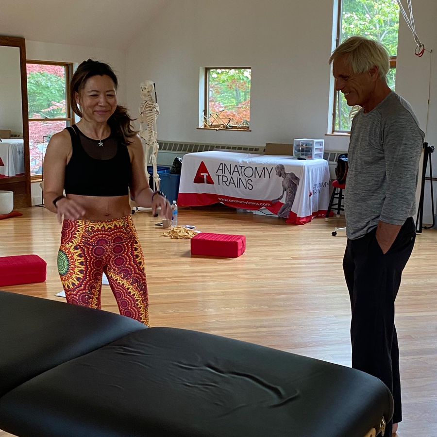Woman in workout clothes and man standing next to massage table in a light-filled studio.