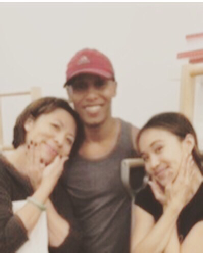 Three people smiling in a dance studio, posing with hands near their faces. One wears a red cap.