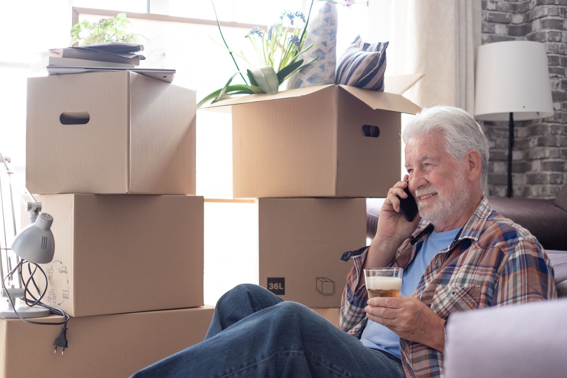 Man sitting among stacked moving boxes, talking on a phone in a bright room