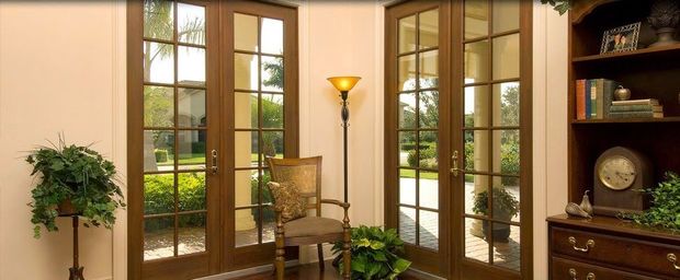 Bright sunroom with French doors, potted plants, a chair, and a wooden bookshelf with clock and decor.
