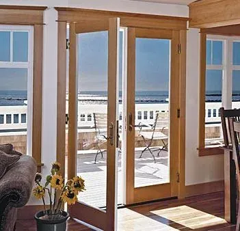 Sunlit living room with wooden French doors opening to an ocean-view patio with table and chairs