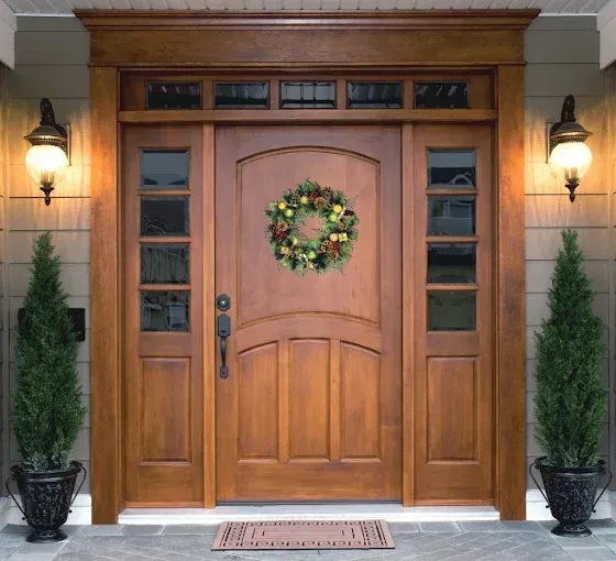 Wooden front door with a wreath, flanked by sconces and potted evergreens on a brick stoop