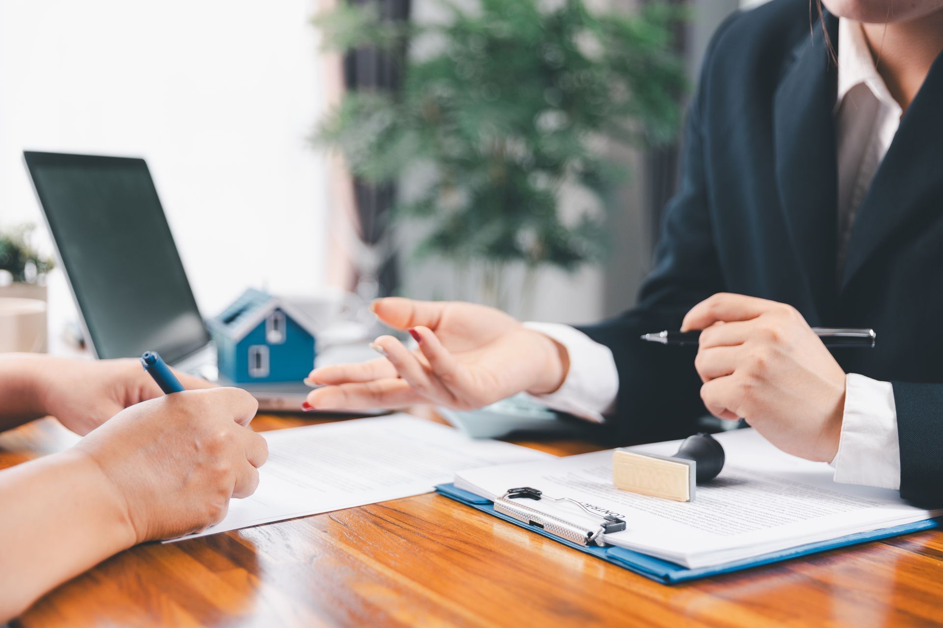 Two people at a table, signing documents. One person gestures, a house model and laptop in the background.