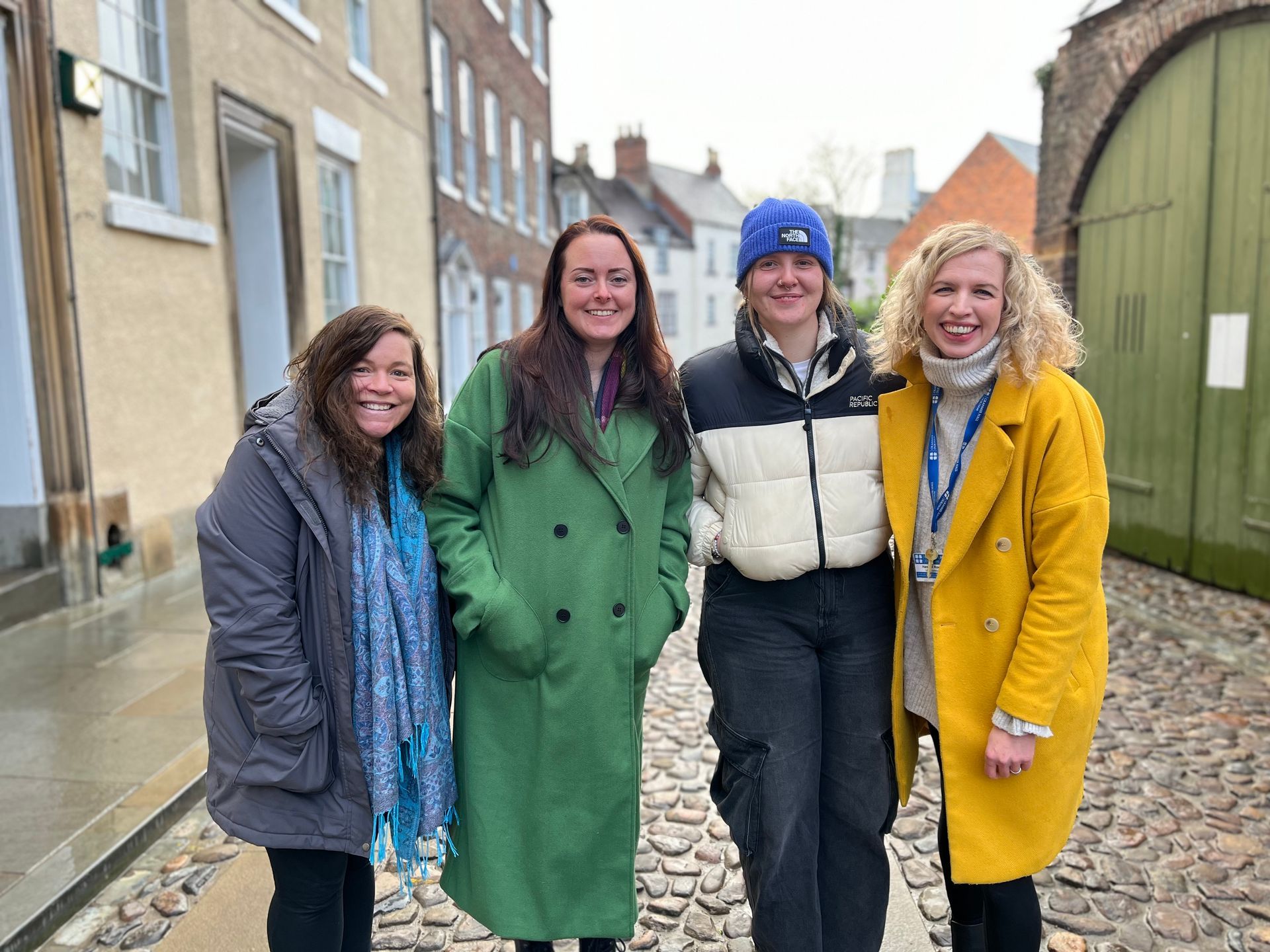 Four people standing together on a street, smiling in colorful coats near brick buildings.