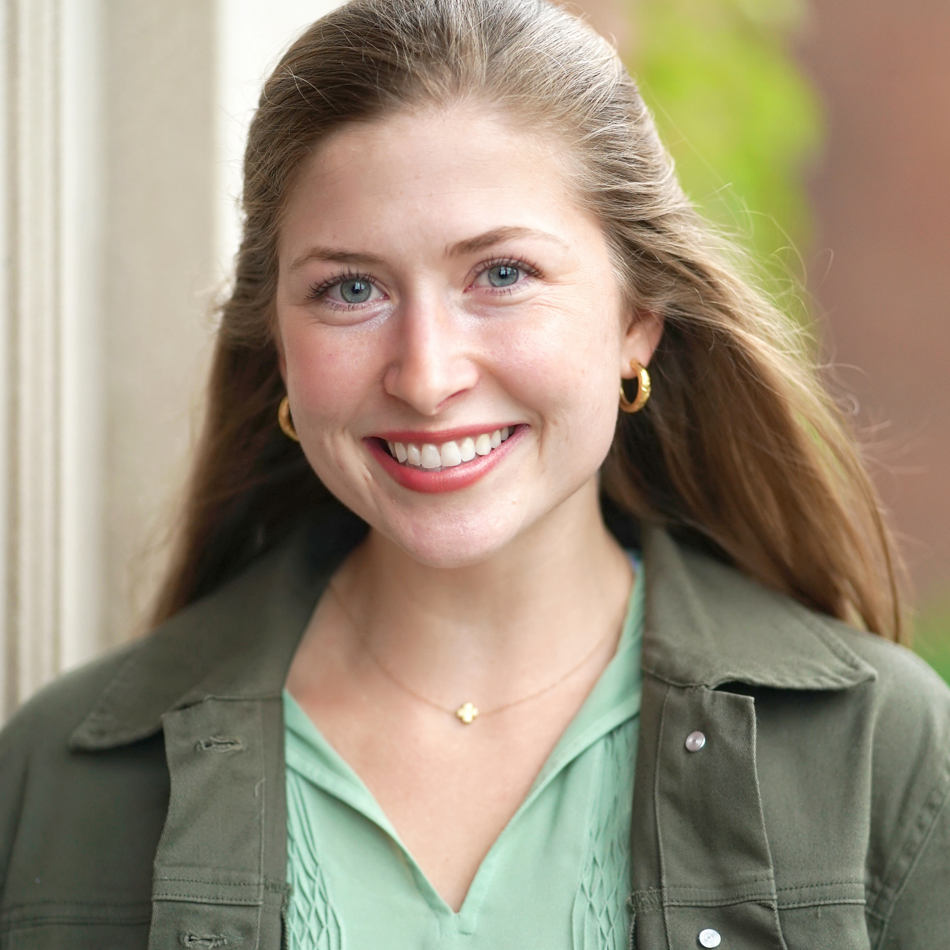 Smiling woman in a green jacket outdoors, with long hair and a softly blurred background