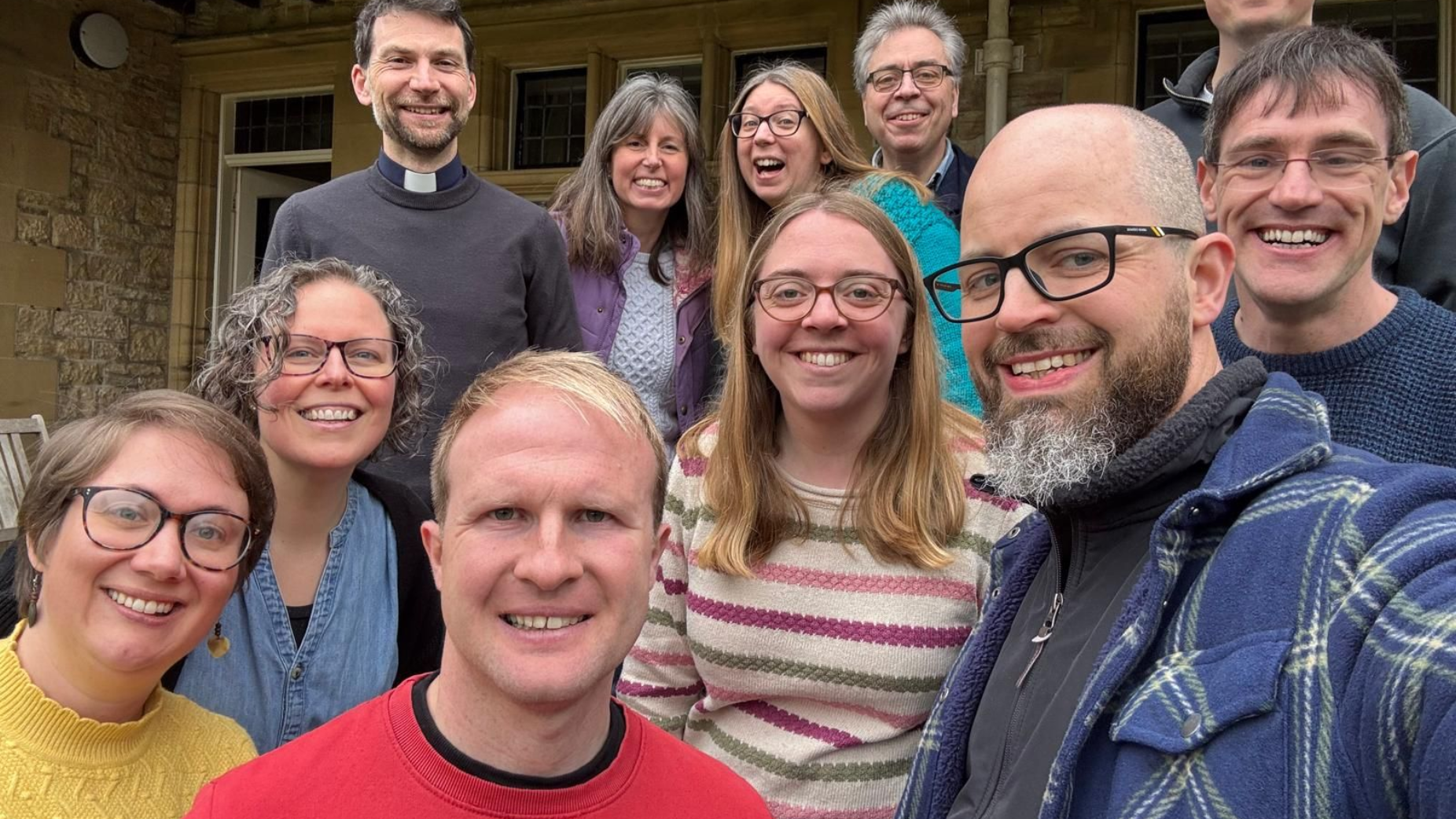 Group of nine people smiling together on a porch, with a tan wall and column behind them.