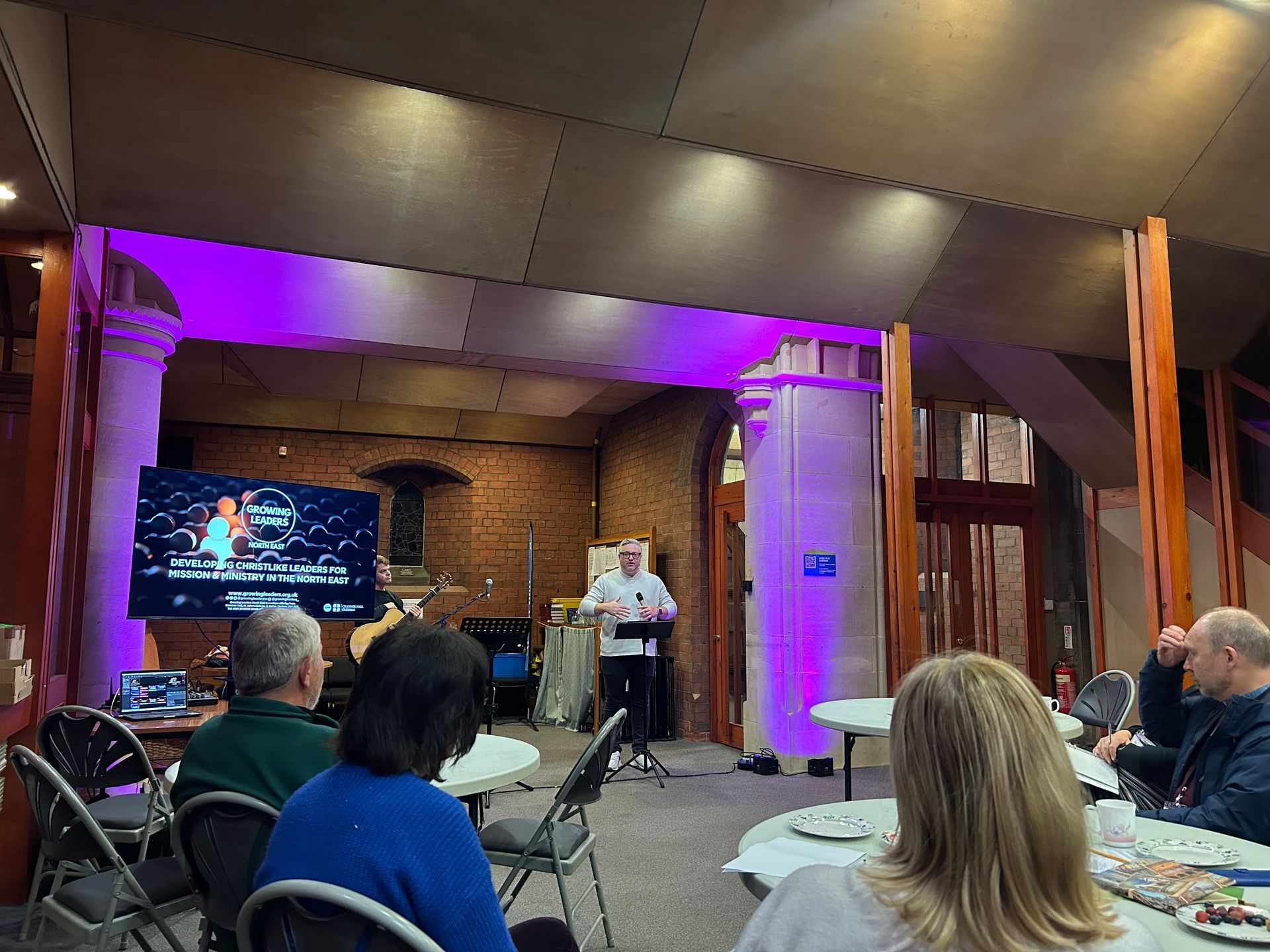 Speaker presenting in a purple-lit hall with seated attendees and a large screen.