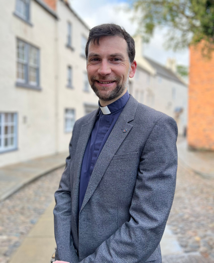 Smiling priest in a patterned gray blazer standing on a cobblestone street with buildings behind him