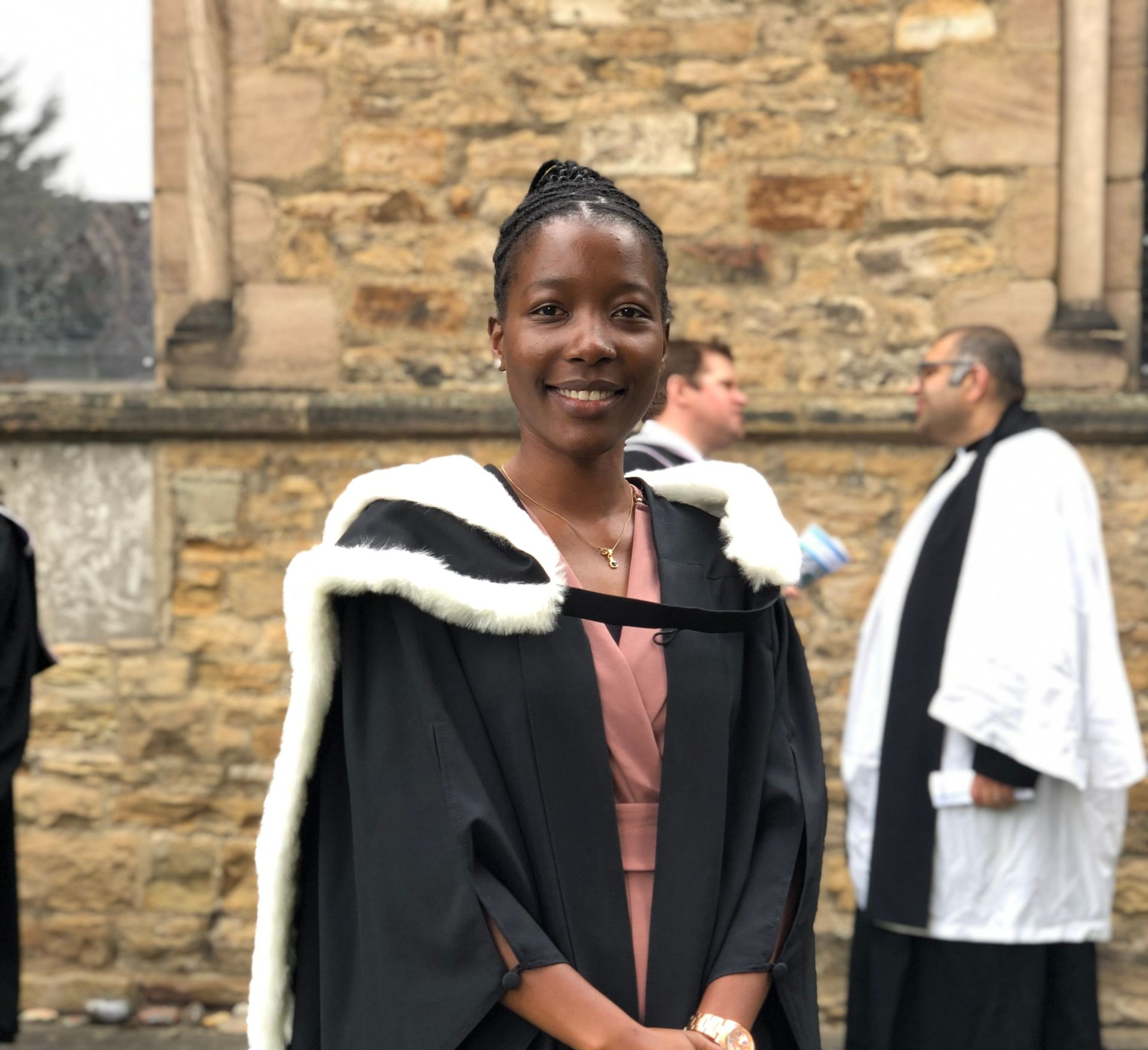 Person in black and white formal attire smiling outdoors in front of a stone building at a graduation ceremony
