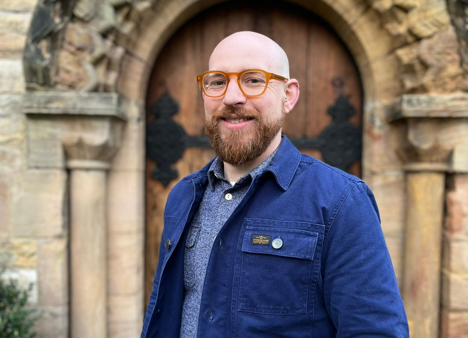 Man in a blue jacket standing in front of a stone archway and wooden door