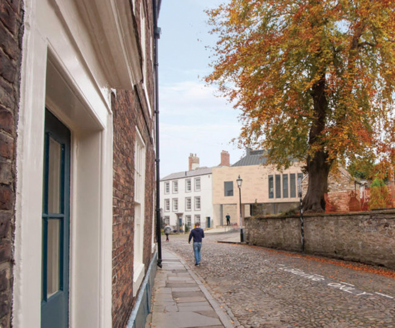 Narrow cobblestone street with white buildings, a lone person walking, and autumn trees by a stone wall