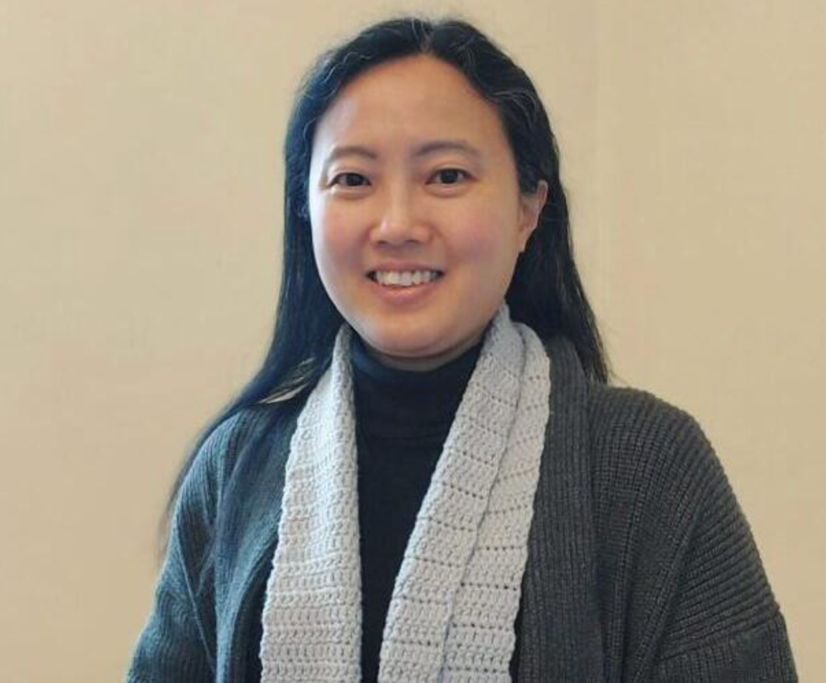 Smiling person in a dark sweater and light scarf against a plain beige background