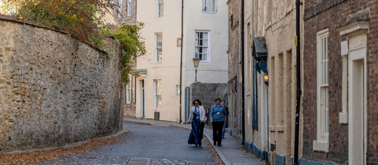 Narrow cobblestone street lined with old buildings, with two people walking beside a bicycle.
