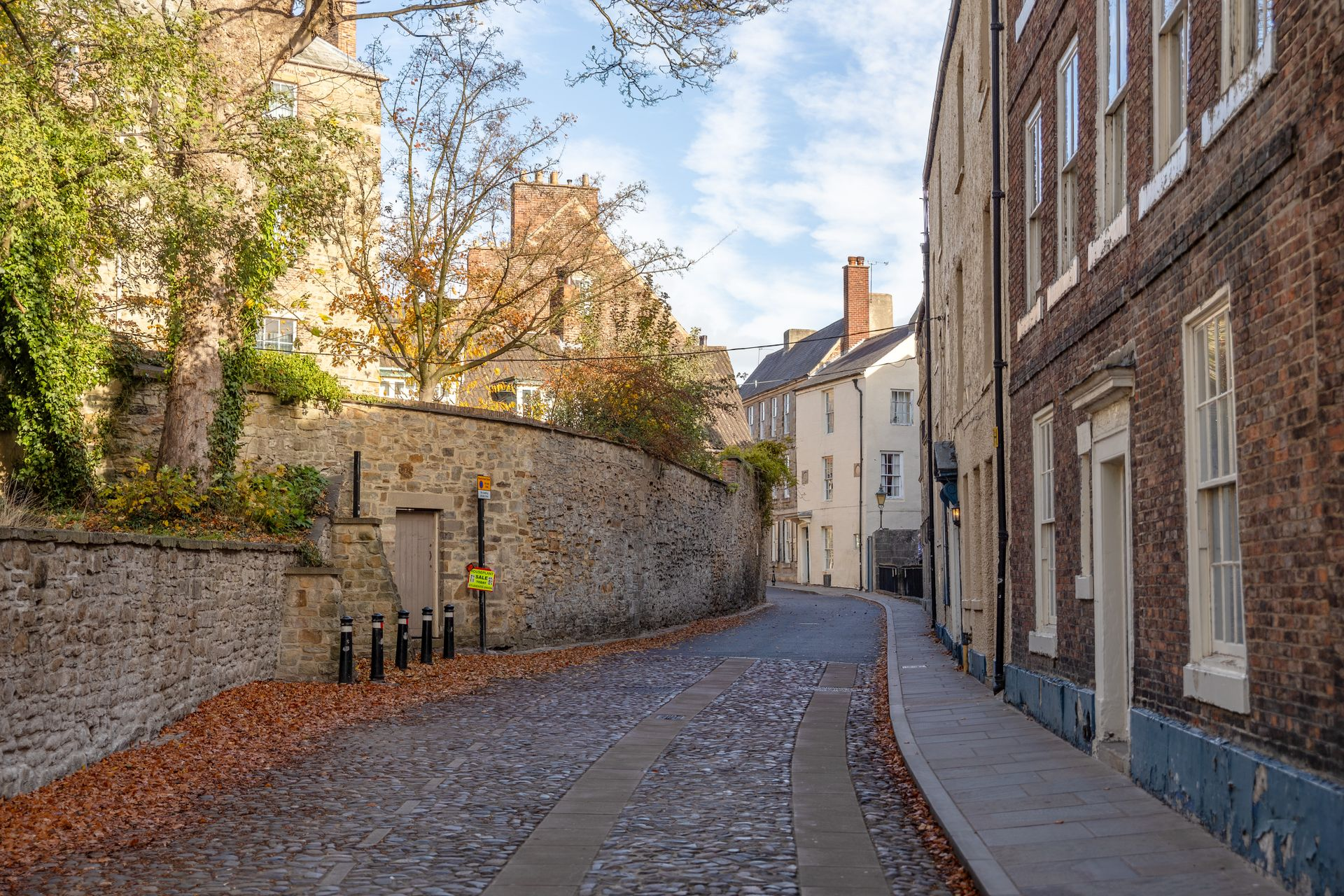 Narrow cobblestone street lined with brick buildings and autumn trees, curving toward a light-colored house.