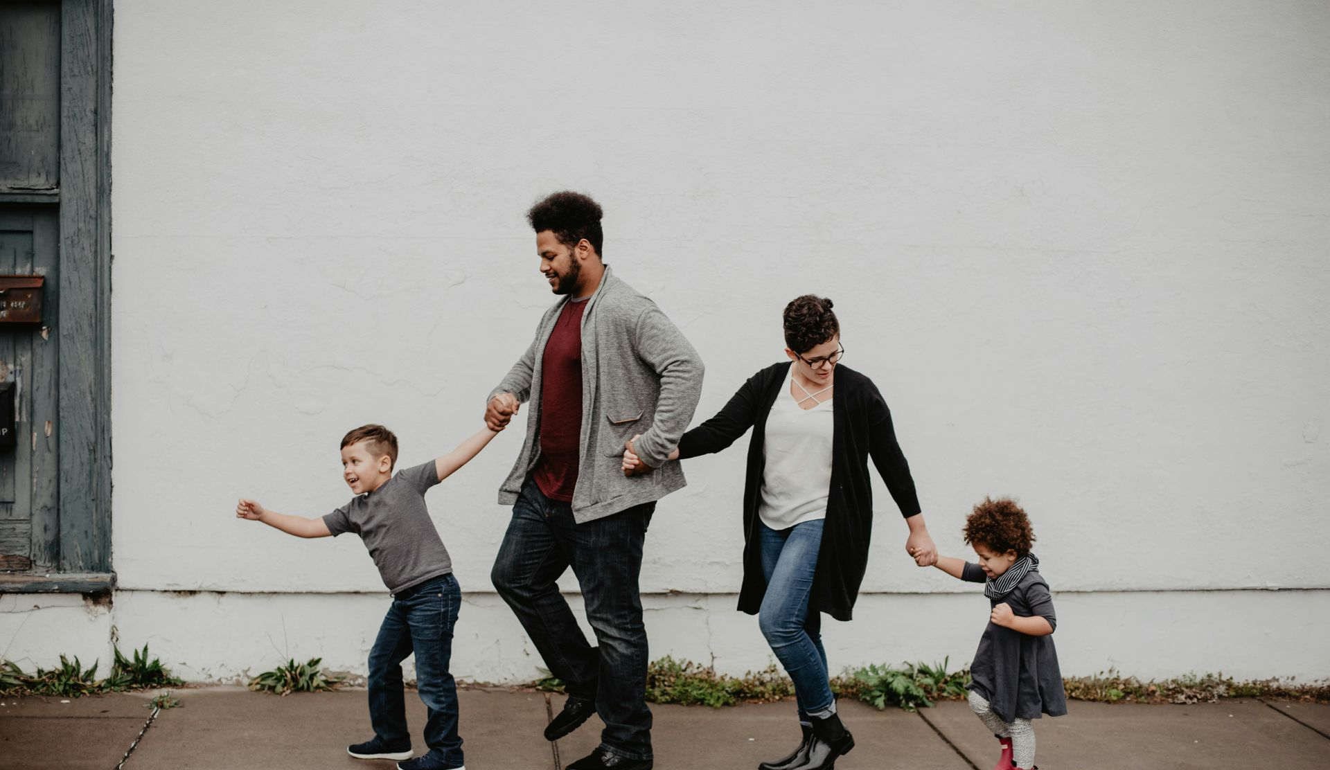 Four people holding hands, walking and smiling along a plain white wall.