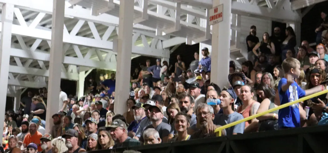 Crowd seated in a tiered arena under white rafters, watching an event.