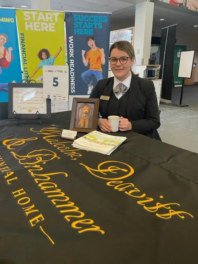 Woman at an event booth in a suit and glasses, standing behind a table with “Princess” banner and promotional materials.