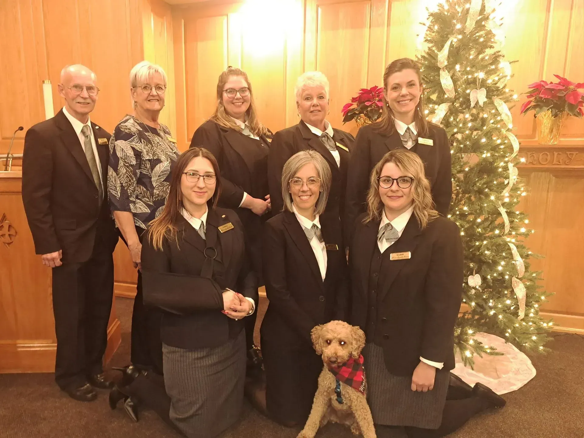 Group posed indoors by a lit Christmas tree, with a small dog in front.