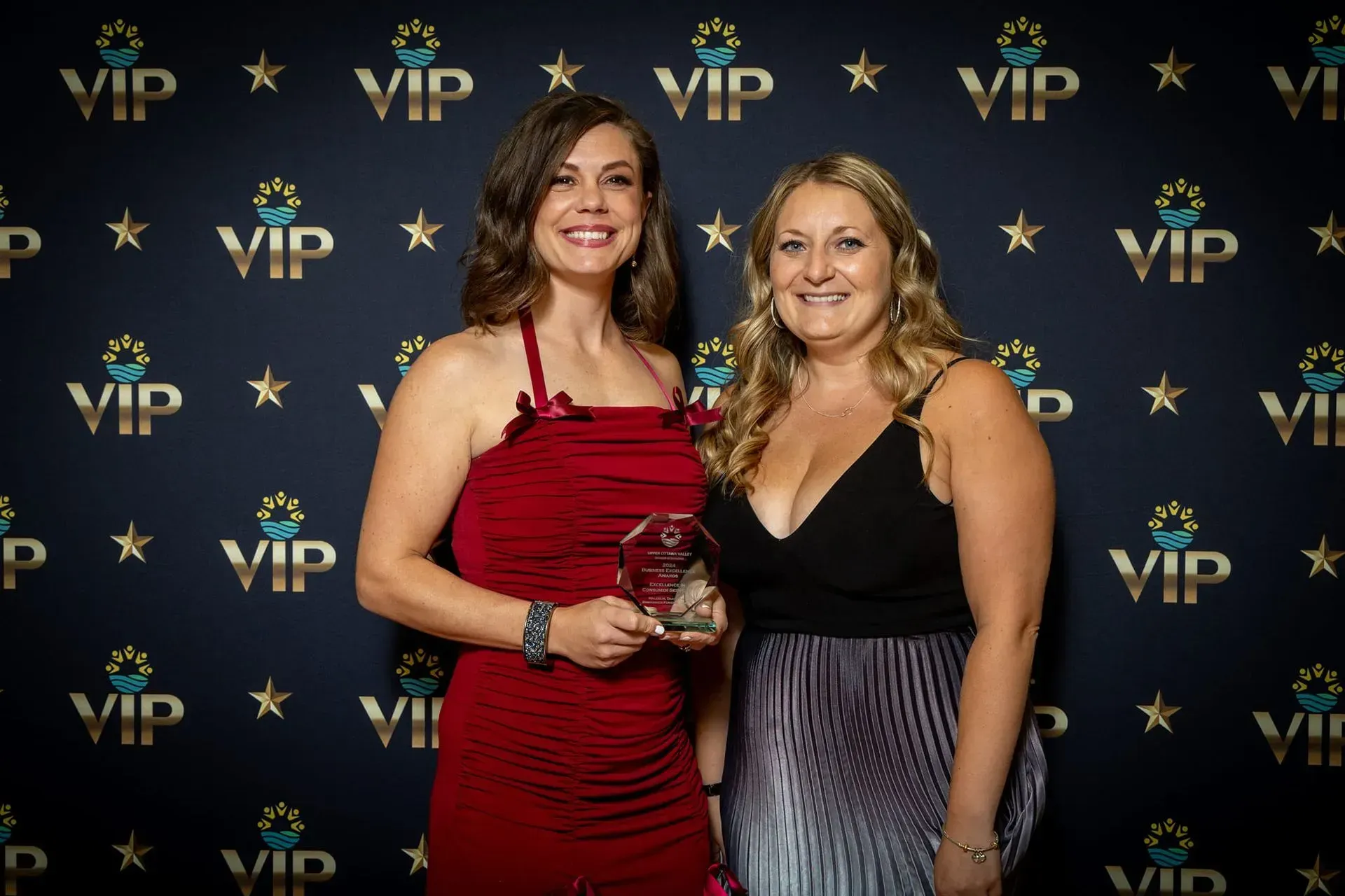 Two women posing at a VIP event backdrop, one holding a glass trophy, both smiling in dresses