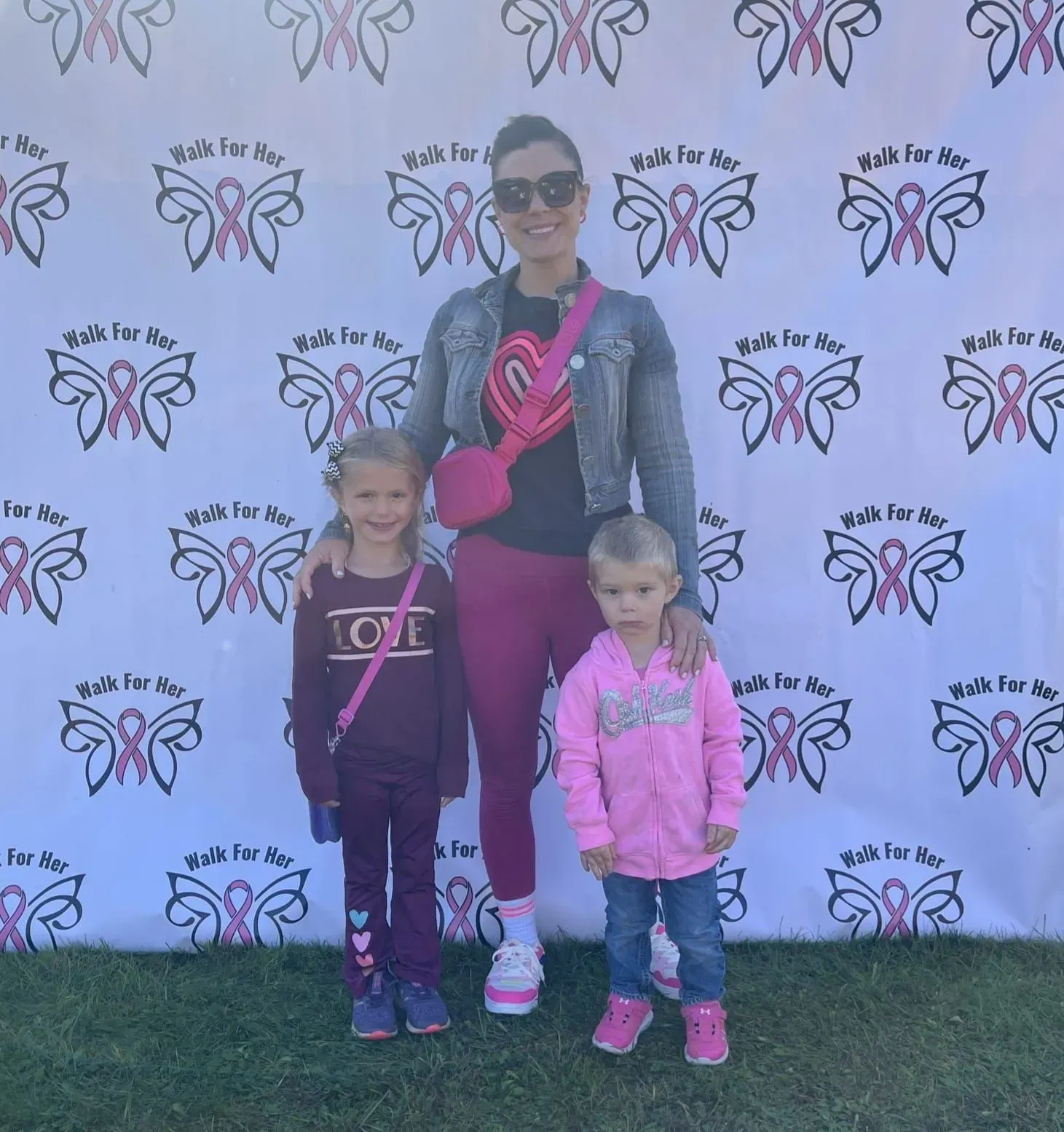 Woman with two children posing at a Walk for a Cure event banner, all wearing pink and sunglasses.