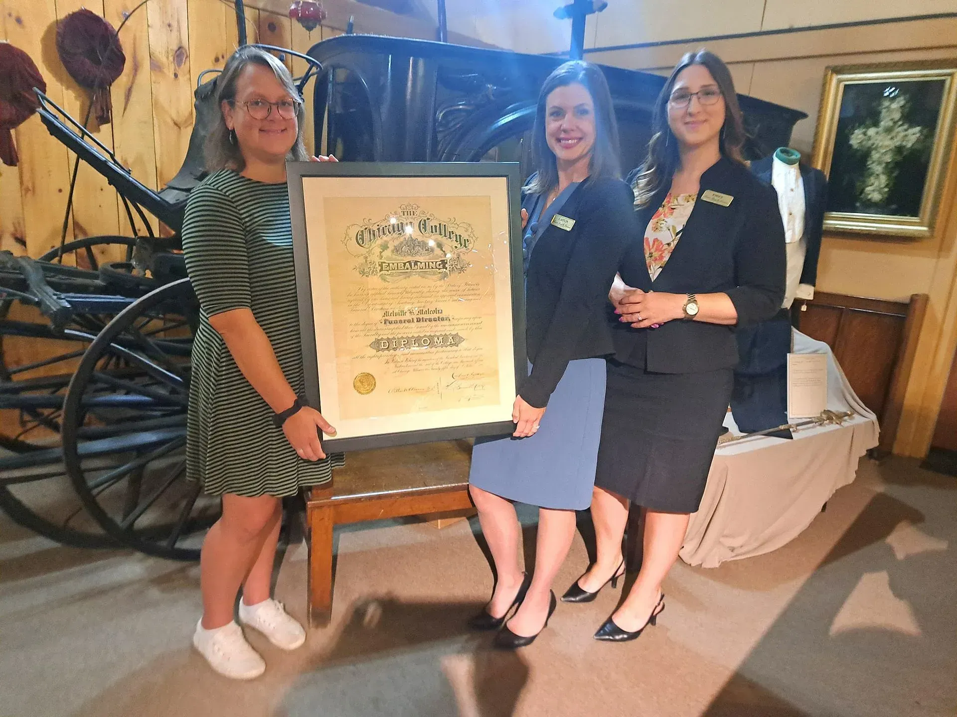 Three women posing with a large framed award plaque in a formal indoor setting