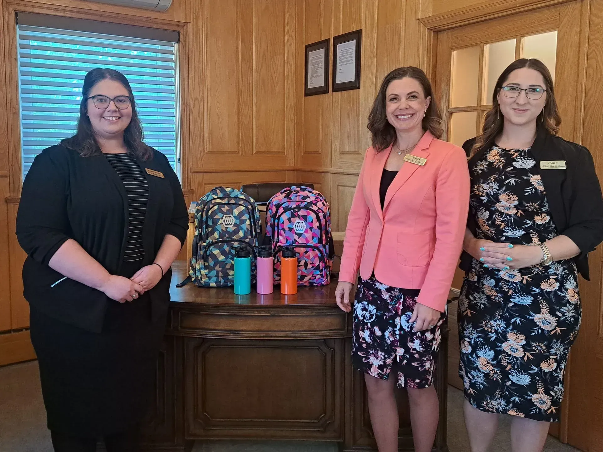 Three women standing indoors beside colorful backpacks and water bottles on a wooden cabinet.