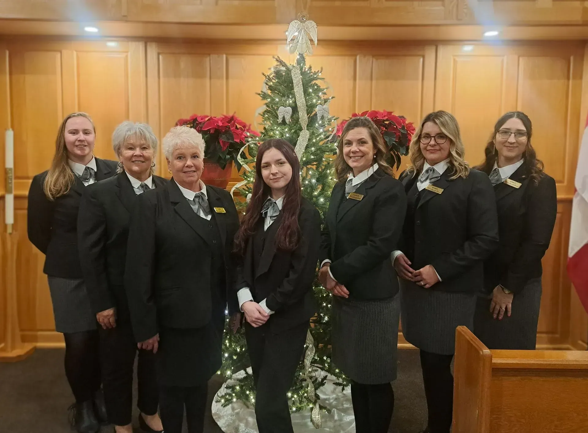 Seven people in black suits stand together in a wood-paneled room beside a decorated Christmas tree.