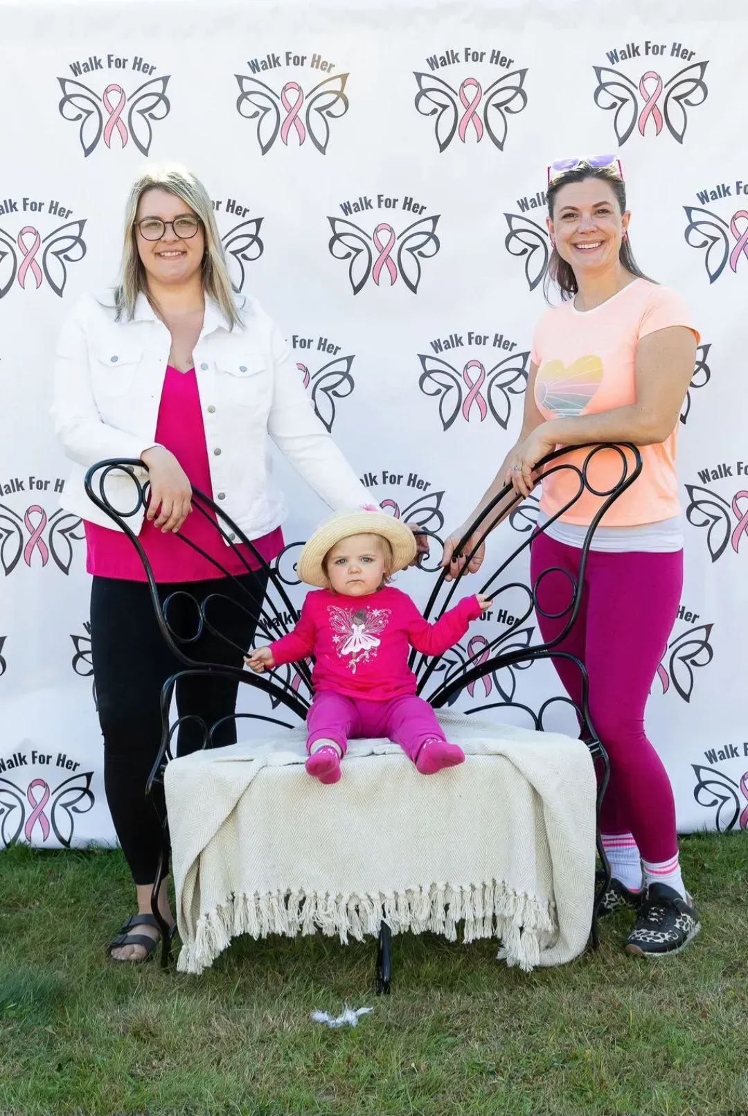 Two women pose with a toddler on a bench in front of a logo backdrop, holding a toy stroller and hoop.