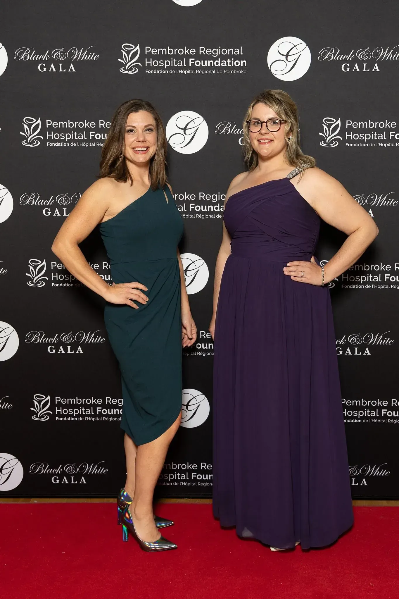 Two women posing on a red carpet in formal dresses before a logo backdrop.