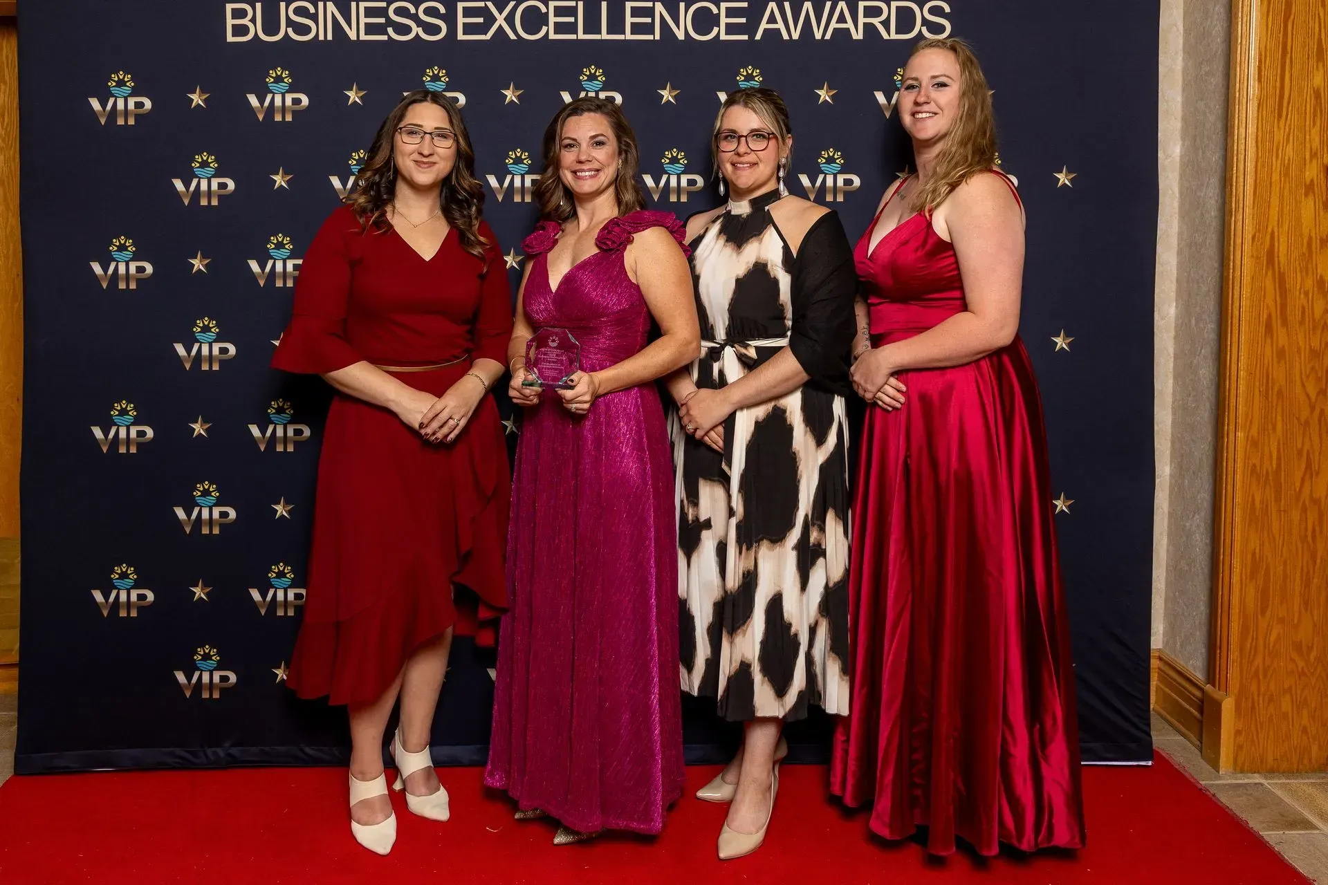 Four people posing on a red carpet in formal dresses at the Business Excellence Awards backdrop