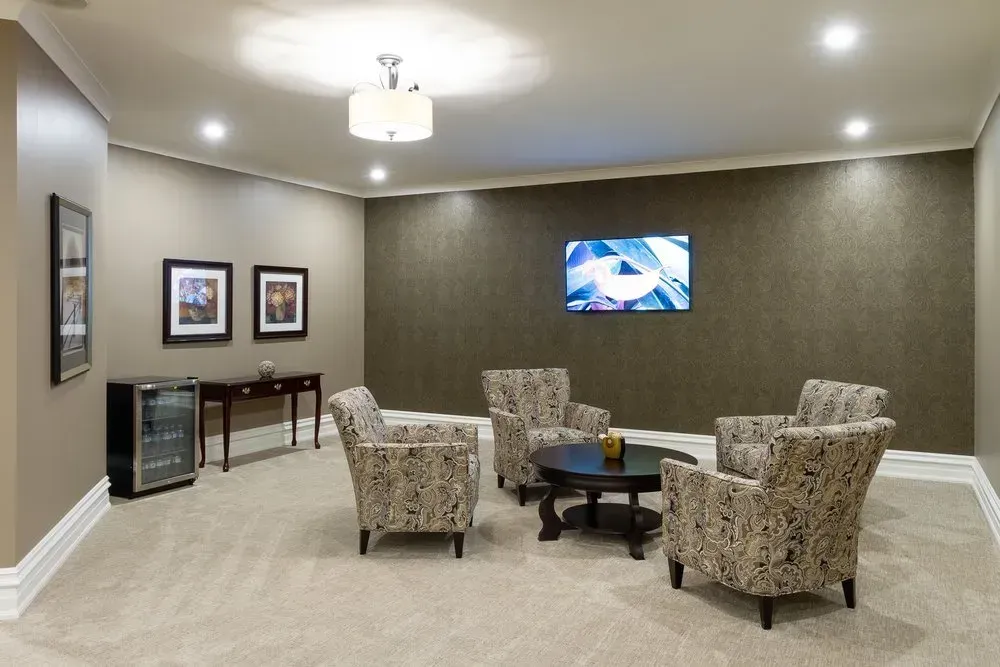 Empty lounge with four patterned chairs, round table, wall-mounted TV, and framed art on beige walls.