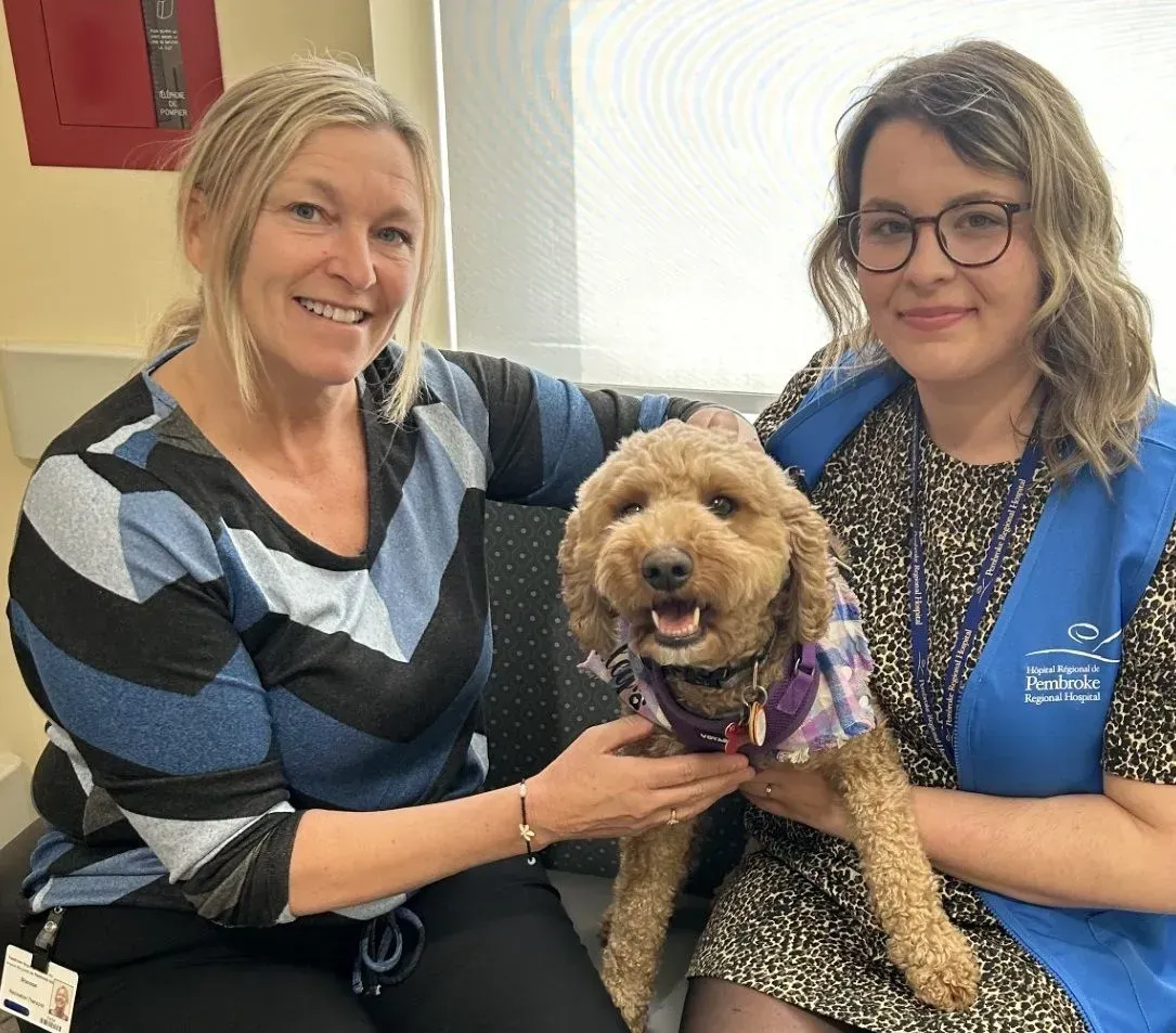 Two women sitting with a small curly dog on a couch, all smiling indoors