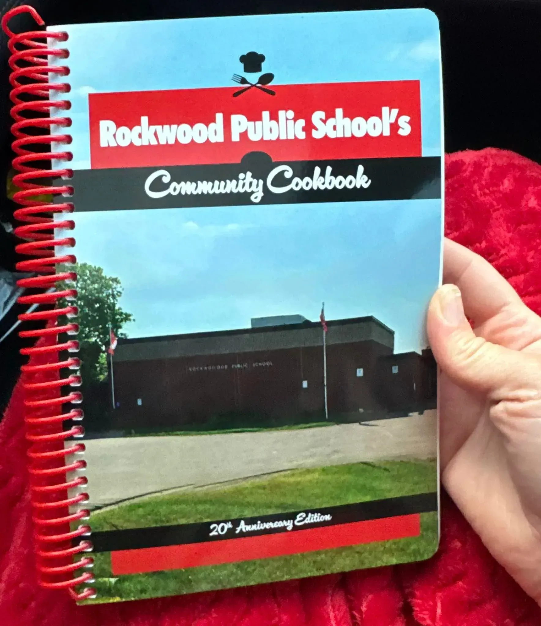 Hand holding a red spiral-bound Redwood Public Schools Community Cookbook with a school photo cover