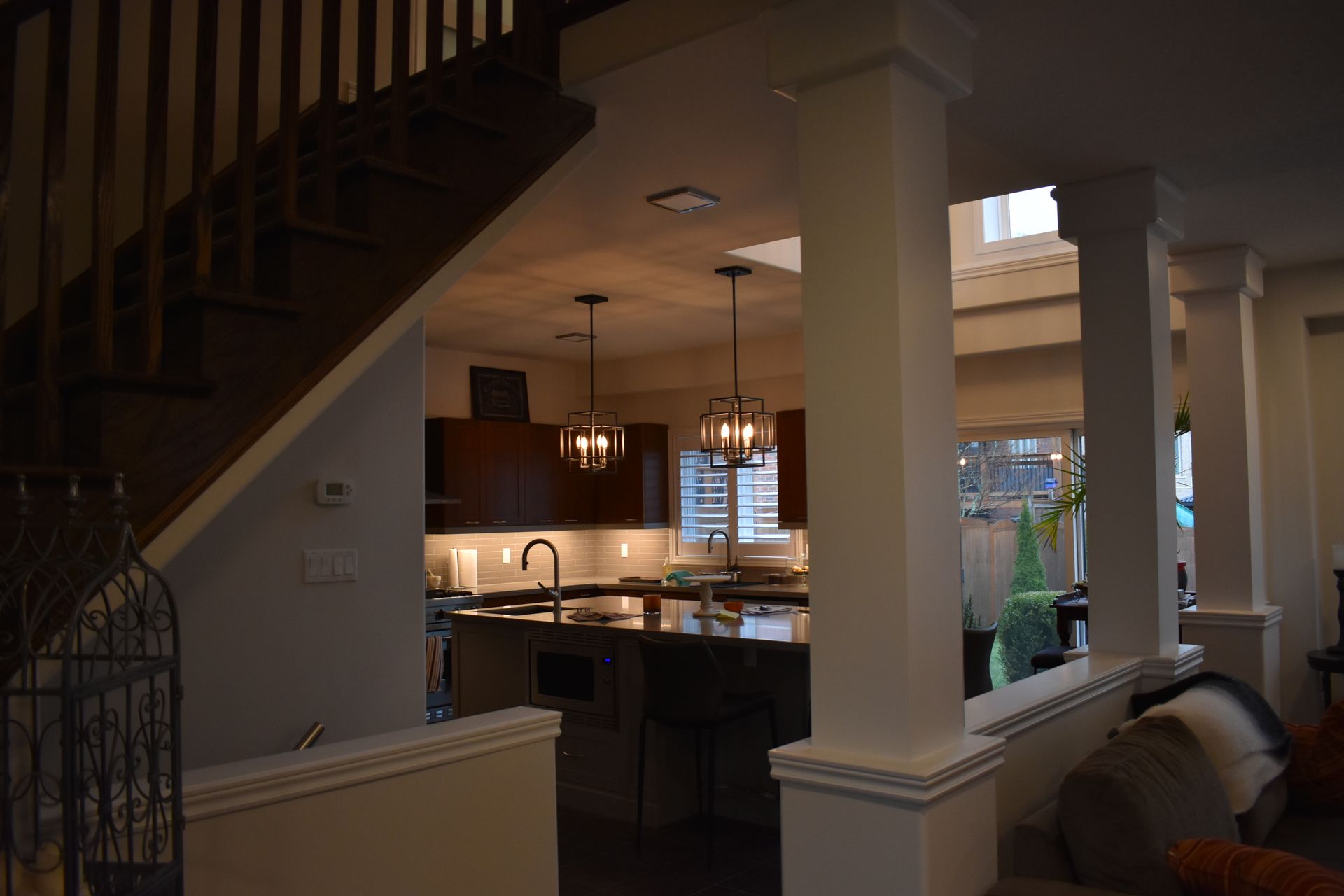 Open kitchen viewed from a living room, with pendant lights, columns, and staircase railing.