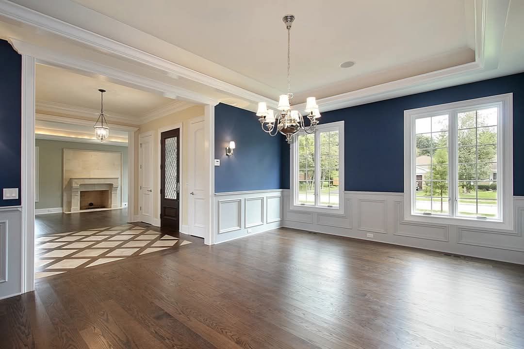 Empty elegant living room with blue walls, hardwood floors, white trim, and large windows.