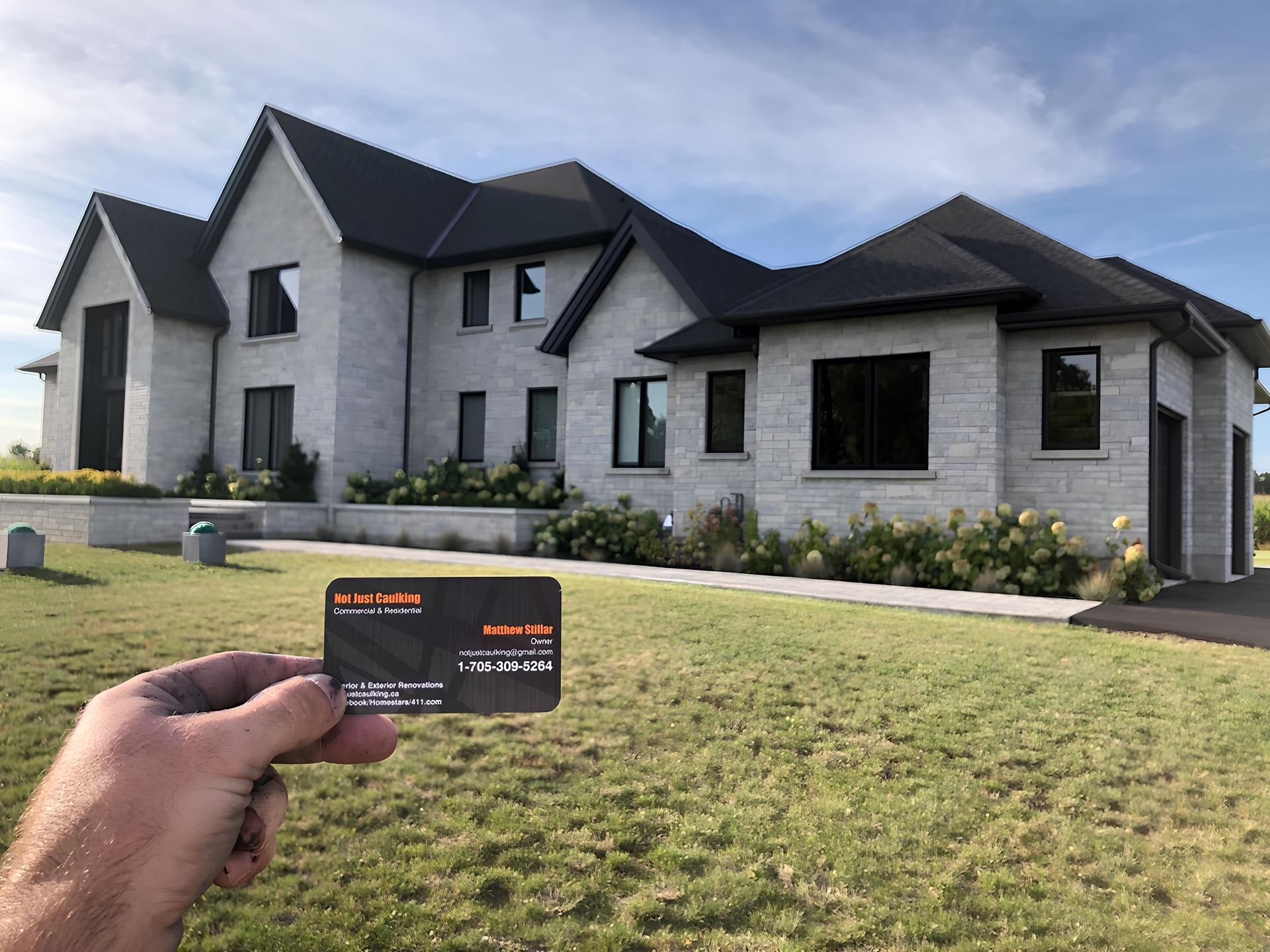 Hand holding business card in front of a light-grey brick house with black roof, green lawn on a sunny day.
