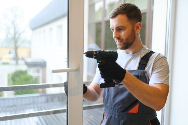 Technician in work overalls and gloves using a drill to install a window latch indoors