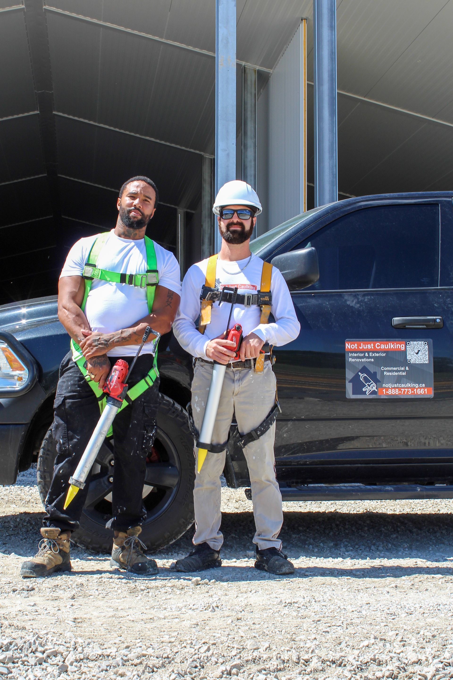 Two men in work harnesses and hard hats stand by a black truck in front of a metal building.