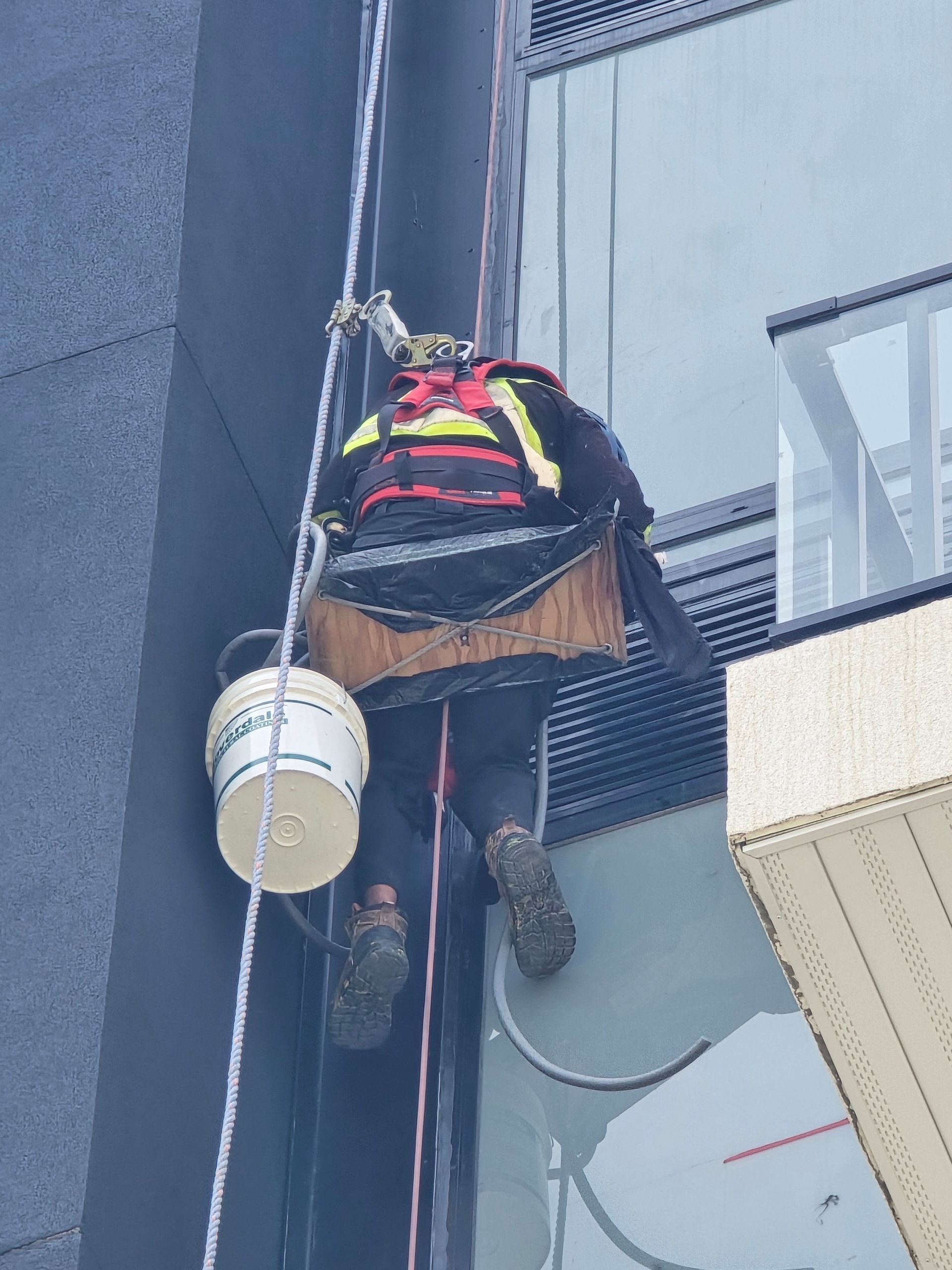 Worker suspended by rope on building facade, wearing a safety harness and helmet, near a white bucket.