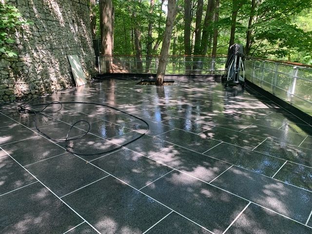Wet, black tiled patio reflecting sunlight. A hose lies on the tiles near a forested area.
