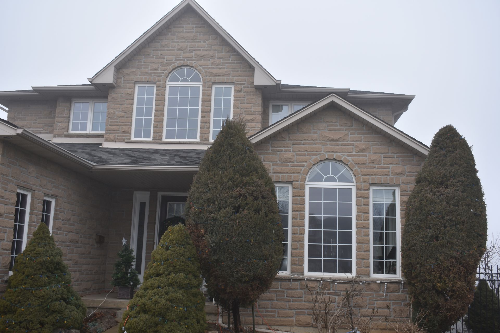 Large brick suburban house with arched windows and tall evergreen shrubs in front