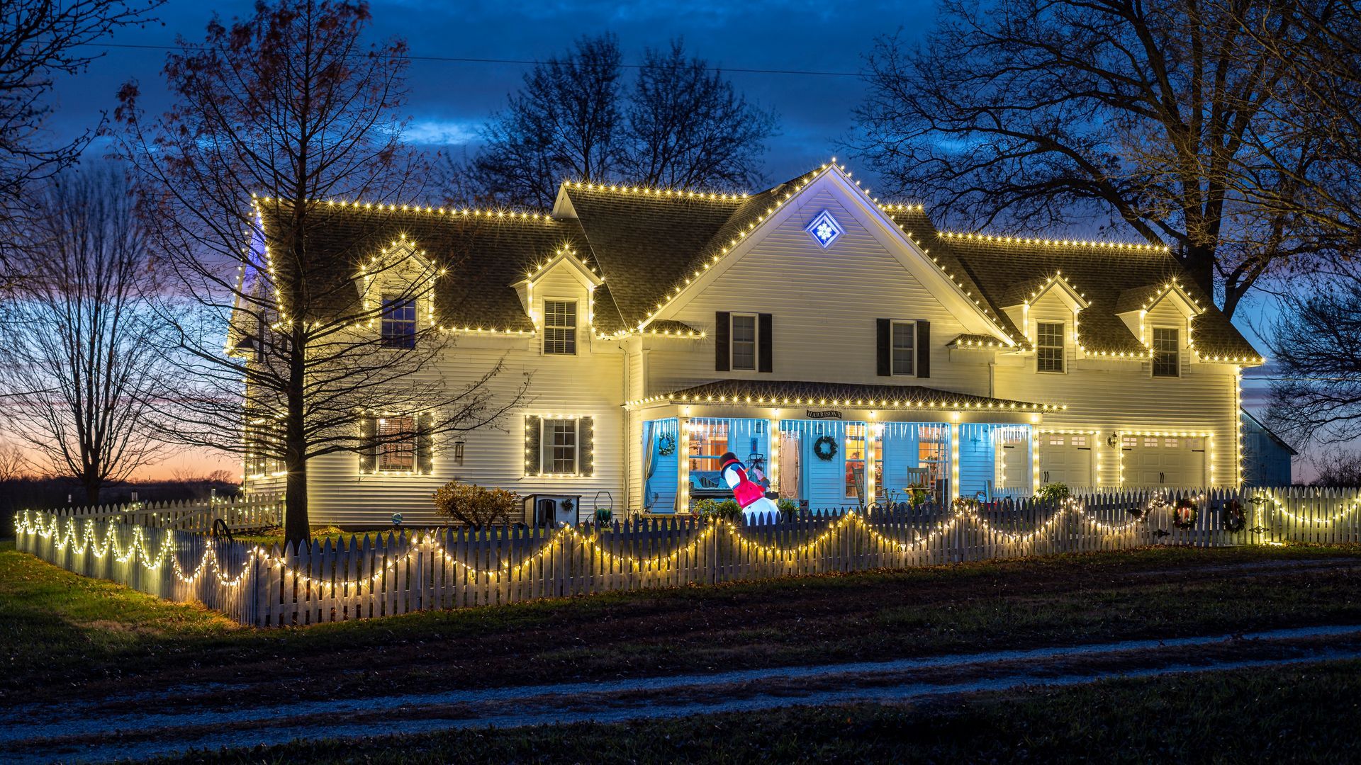 Large house at dusk outlined with warm white Christmas lights, with blue-lit windows and a snowy yard